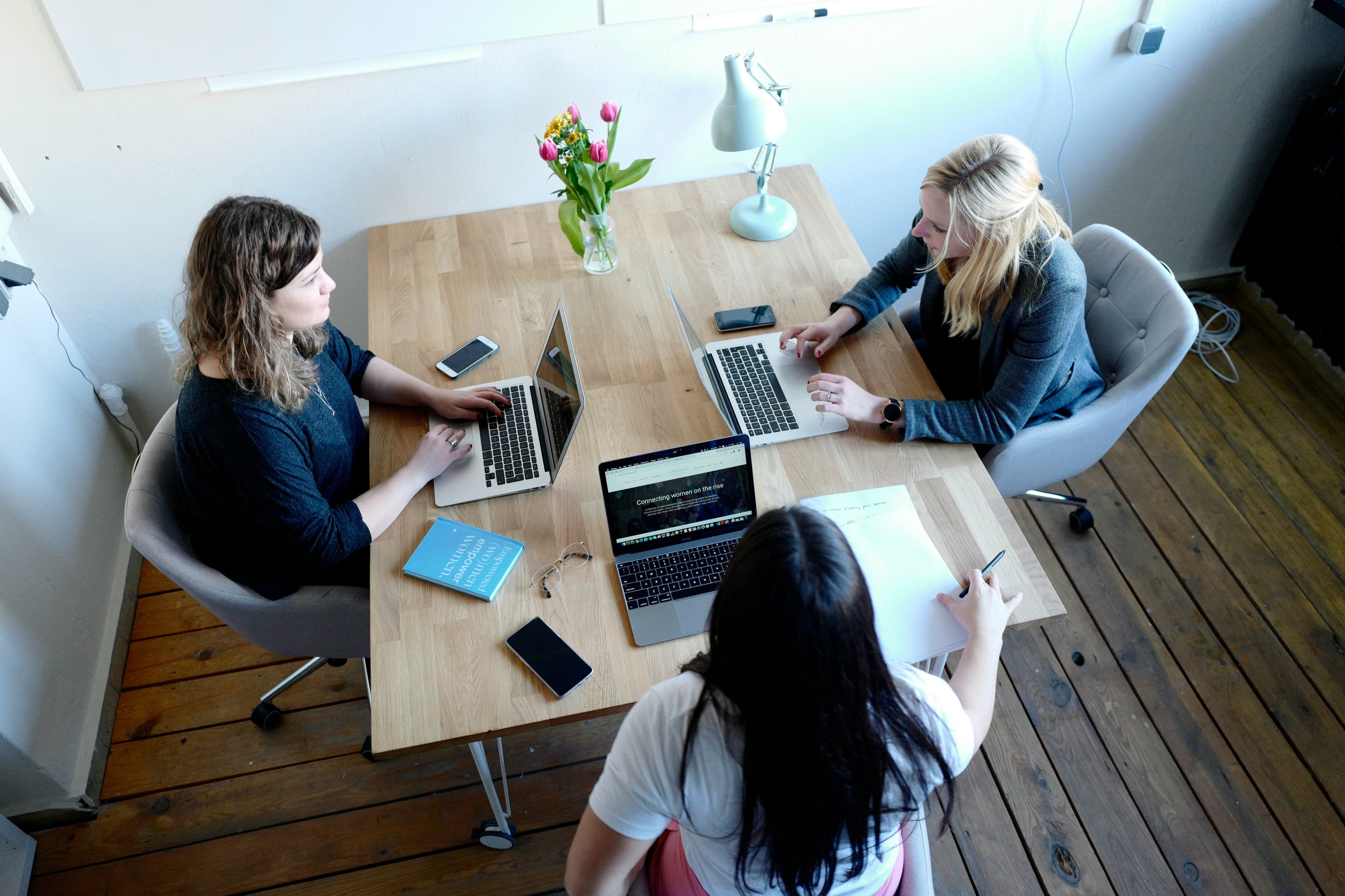 Three people working on laptops at a wooden table with a vase of flowers in the center.